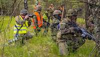 Infanteristen hocken im Gras und bereiten sich zum Sturm auf den Feind vor, darunter ein Fotograf.