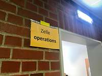 Callcenter Reservisten: Zelle Operations Ein gelbes Schild mit der Aufschrift "Zelle Operations" an einer Backsteinwand, daneben eine offene Tür.