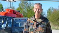 Ein Soldat im Feldanzug steht im Freien vor einem Hubschrauber und lächelt.