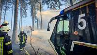 Ein Feuerwehrfahrzeug in einem Waldgebiet, daneben Feuerwehrleute