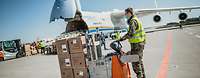 Ein Soldat mit Schutzmaske transportiert Kartonagen mit einem Hubwagen, im Hintergrund steht das Frachtflugzeug Antonow