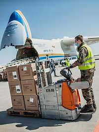 Ein Soldat mit Schutzmaske transportiert Kartonagen mit einem Hubwagen, im Hintergrund steht das Frachtflugzeug Antonow