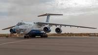 Die Iljuschin IL-76 steht auf ihrer Parkposition. Ein Flughafenmitarbeiter positioniert die Antiwegrollhölzer am Fahrwerk.