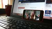 Ein aufgeklappter Laptop, darauf steht quer ein Handy. Auf dem Handy ist eine junge Frau in einer Videkonferenz zu sehen.