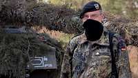 Ein Soldat mit schwarzem Mundschutz und schwarzem Barett steht vor einem Kampfpanzer.