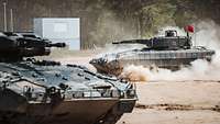 Zwei Panzer fahren in einem großen Bogen über sandiges Gelände und wirbeln Staub auf.