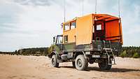 Ein Lkw mit einem orangefarbenen Zeltaufbau steht auf einer sandigen Fläche in der Sonne.
