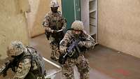 Zwei Soldaten stürmen bewaffnet einen Raum. Ein dritter Soldat im Hintergrund bewertet das Handeln.