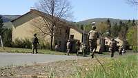 Soldaten mit Helm, Schutzweste und Gewehr (von hinten gesehen) gehen im urbanene Gelände eine Straße entlang