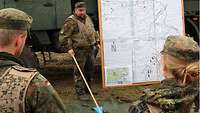 Ein Soldat mit Zeigestab in der Hand steht neben einer großen Übungsskizze, zwei Soldaten (von hinten gesehen) hören ihm zu