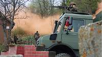 Ein Soldat bedient stehend in einem Lkw-Unimog ein Maschinengewehr auf einer Ringlafette, davor Mauerreste, dahinter roter Rauch