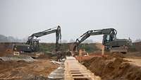 Zwei Bagger arbeiten an einer Baustelle. Drei Soldaten stehen in der Grube. 