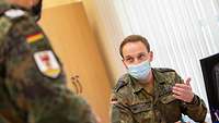 Ein Soldat sitzt mit einem Mundschutz am Schreibtisch und spricht mit einem anderen Soldaten. 