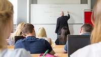 Blick in einen Hörsaal: im Vordergrund Studierende, im Hintergrund eine Tafel, auf die eine Person schreibt