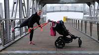 Eine Frau beim Sport mit einem Gymnastikband vor einem Kinderwagen, der auf einer großen Brücke steht