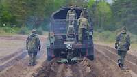 Zwei Soldaten laufen über aufgewühlte Erde rechts und links hinter dem Lkw mit Anhänger her.