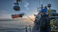 Auf dem Achterschiff eines kleinen grauen Kriegsschiffs steht ein Marinesoldat in dunkelblauer Arbeituniform.