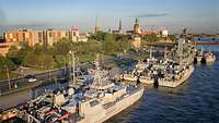 Luftaufnahme mehrerer kleiner, grauer Kriegsschiffe, die in einem Hafen liegen; im Hintergrund Kirchtürme einer Stadt.
