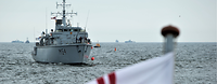 Open Spirit Ein kleines graues Kriegsschiff in See, davor eine weiße Flagge mit einem roten Kreuz mit doppelten Linien.