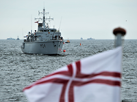Open Spirit Ein kleines graues Kriegsschiff in See, davor eine weiße Flagge mit einem roten Kreuz mit doppelten Linien.