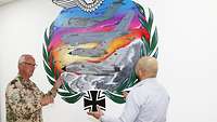 Zwei Männer, einer in Uniform, einer in Zivil, stehen einander gegenüber und betrachten ein Bild an der Wand
