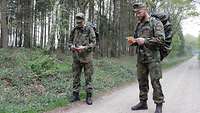 Zwei Soldaten mit Gepäck stehen auf einem Waldweg, einer mit Kompass, der andere mit Meldezettel.