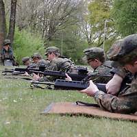 Soldaten liegen in schräger Reihe auf Kokosmatten und zerlegen Gewehre. Der Ausbilder beobachtet sie