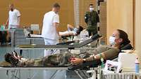 Eine Soldatin liegt auf einer Liege und spendet Blut, im Hintergrund zwei Sanitäter und ein Soldat.