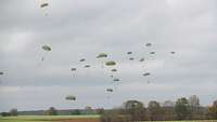 Am Himmel gleiten Fallschirmjäger an ihren Schirmen zu Boden.