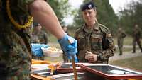 Ein Soldat schöpft Nudeln aus einem Wärmebehälter, eine Soldatin davor wartet auf ihr Essen.