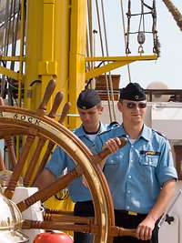 Vier Marinesoldaten in blauer Arbeitsuniform halten ein großes, rundes hölzernes Schiffsruder.