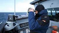 Ein Marinesoldat in dunkelblauer Uniform steht draußen neben der Brücke eines Schiffs und blickt durch ein Fernglas.