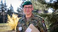 Ein Soldat steht mit einer Unterlagenmappe im Arm neben einem Baum. 