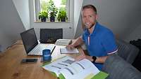 Ein blonder Mann im blauen Poloshirt am Tisch mit Block, Stift, Laptop und Lernunterlagen.