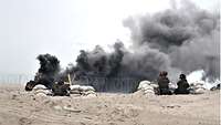 Zwei Soldaten in Flecktarn am Strand, in der Mitte des Bildes schwarze Rauchschwaden.