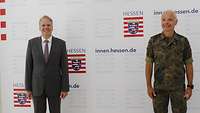 Ein Mann im Anzug und ein Soldat stehen vor einer Wand.