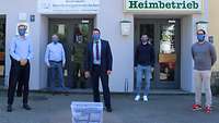 Vor einem Gebäude stehen mehrere Männer in Uniform und Zivil um eine Kiste mit Schutmasken.
