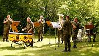 Fünf Bläser und ein Paukenspieler in Uniform stehen auf einer Wiese und musizieren im Sonnenschein.