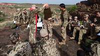 Soldaten sitzen auf Steinen, ein Pfarrer in Uniform legt seine Hand auf die Stirn eines Soldaten.