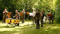 Fünf Bläser und ein Paukenspieler stehen auf einer Wiese und musizieren im Sonnenschein.
