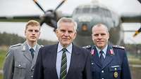 Ein älterer Mann und zwei Soldaten stehen zusammen vor einem Militärflugzeug