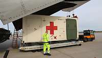 Ein Container mit einem roten Kreuz wird mit dem Kran auf ein Transportfahrzeug gestellt