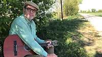 Gunther Wiendl sitzt mit seiner Gitarre im Schatten eines blühenden Busches an einem Wegrand.