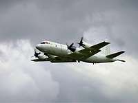 MPA P-3C Orion Ein hellgraues viermotoriges Propellerflugzeug im Flug vor grauem Himmel.