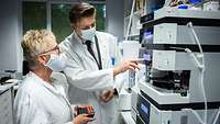 Dr. Koller und Dr. John stehen im Labor vor einem Analysegerät