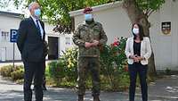 Eine weibliche und zwei männliche Personen, eine davon in Uniform, stehen im Freien mit Abstand und Mund-Nase-Bedeckung