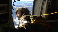 Ein Soldat mit Helm und Fallschirm schaut aus der geöffneten Seitentür eines Flugzeuges.