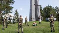 Soldaten und ein Militärpfarrer stehen weit auseinander an einem Hügel vor einem hohen Ehrenmal.