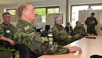 An einem breiten Konferenztisch sitzen drei Soldaten in Abständen voneinander und schauen nach vorn.