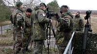 Fünf Soldaten stehen am Rande eines Übungsgeländes, zwei stellen eine aufgebaute Videokamera ein.